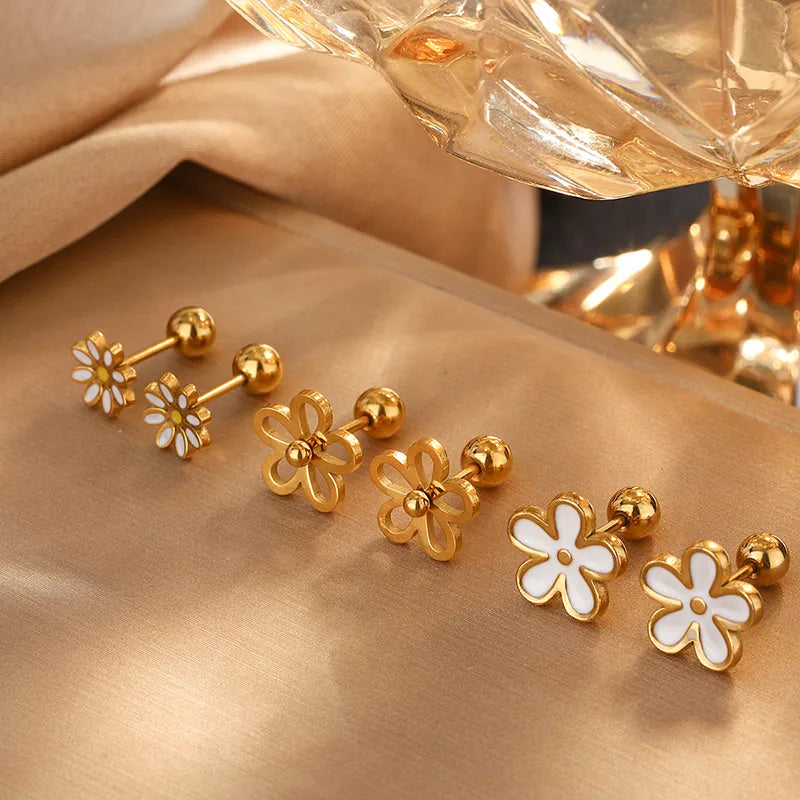 Gold and white flower-shaped earrings on a gold surface with a perfume bottle in the background.