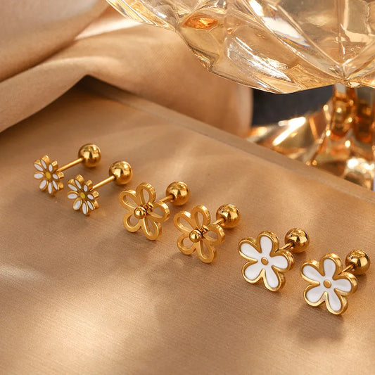 Gold and white flower-shaped earrings on a gold surface with a perfume bottle in the background.